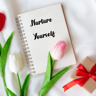 Nurture Yourself written on a white cover of a spiral bound journal. Roses and a gift box a spread around