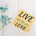 Grey wooden background with blue flowers on the left, and a sticky note with the words, Live the life you love. Get unstuck